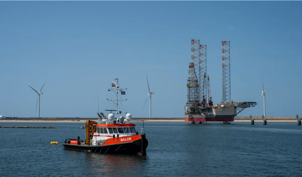 A tugboat named 'Baloe' in the foreground, navigating calm waters, with an offshore oil rig and wind turbines in the background.